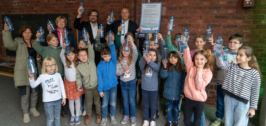 Schoolchildren and four adults stand around a drinking water dispenser.